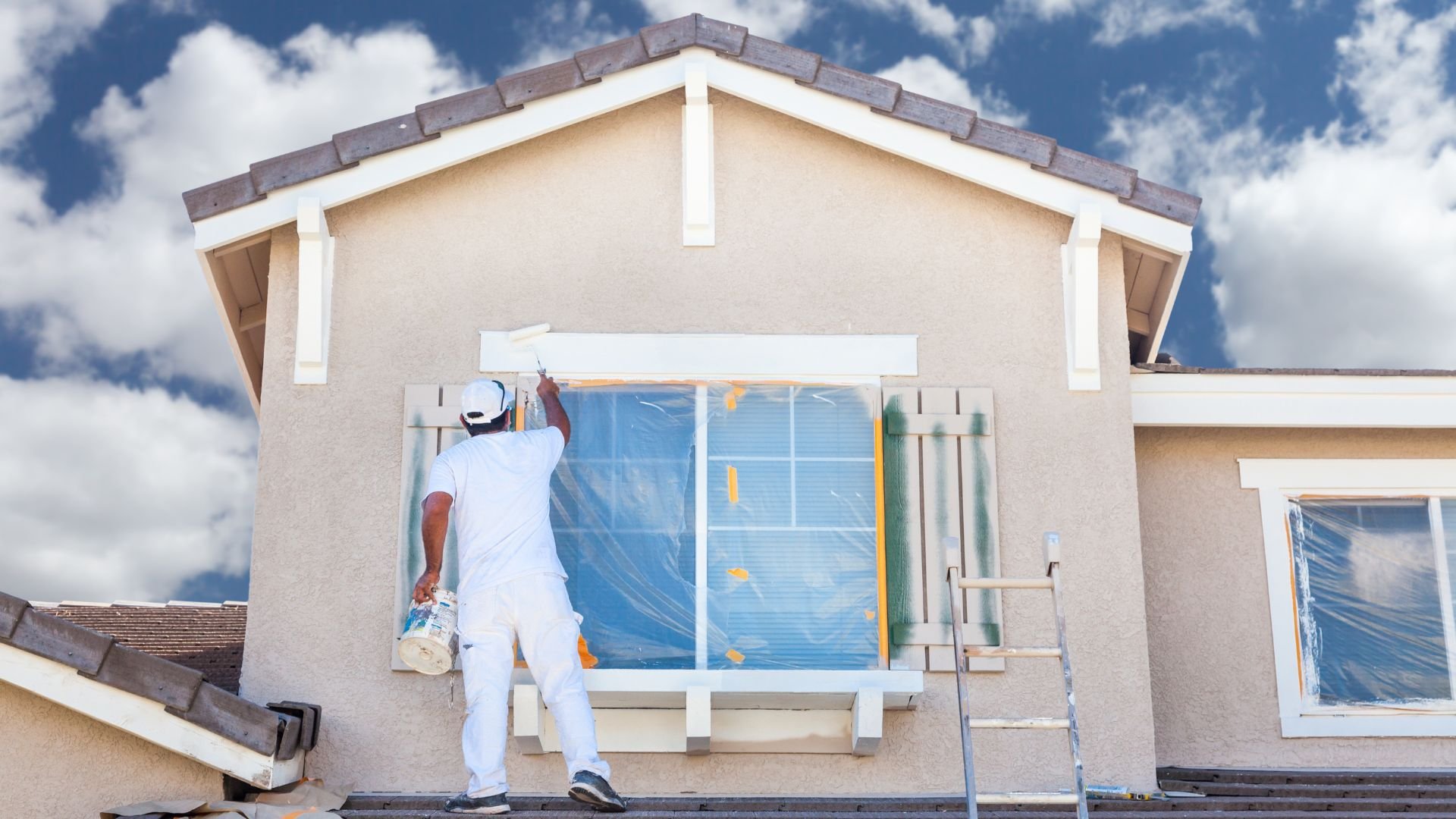 Painter in white outfit painting exterior of house with blue sky backdrop
